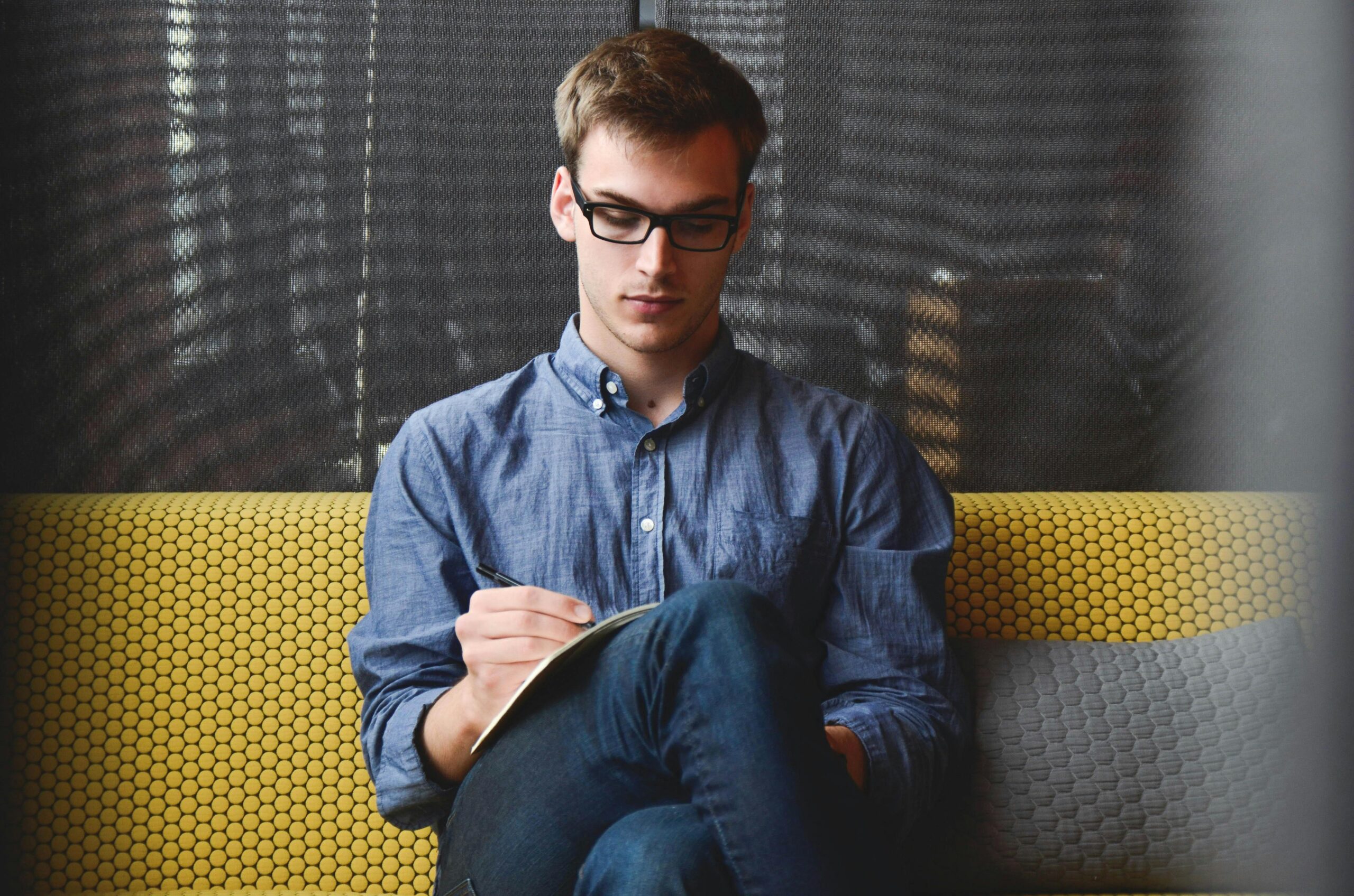 entrepreneur-startup-start-up-man-39866-39866 A young man in glasses writes in a notebook while sitting on a stylish couch indoors.