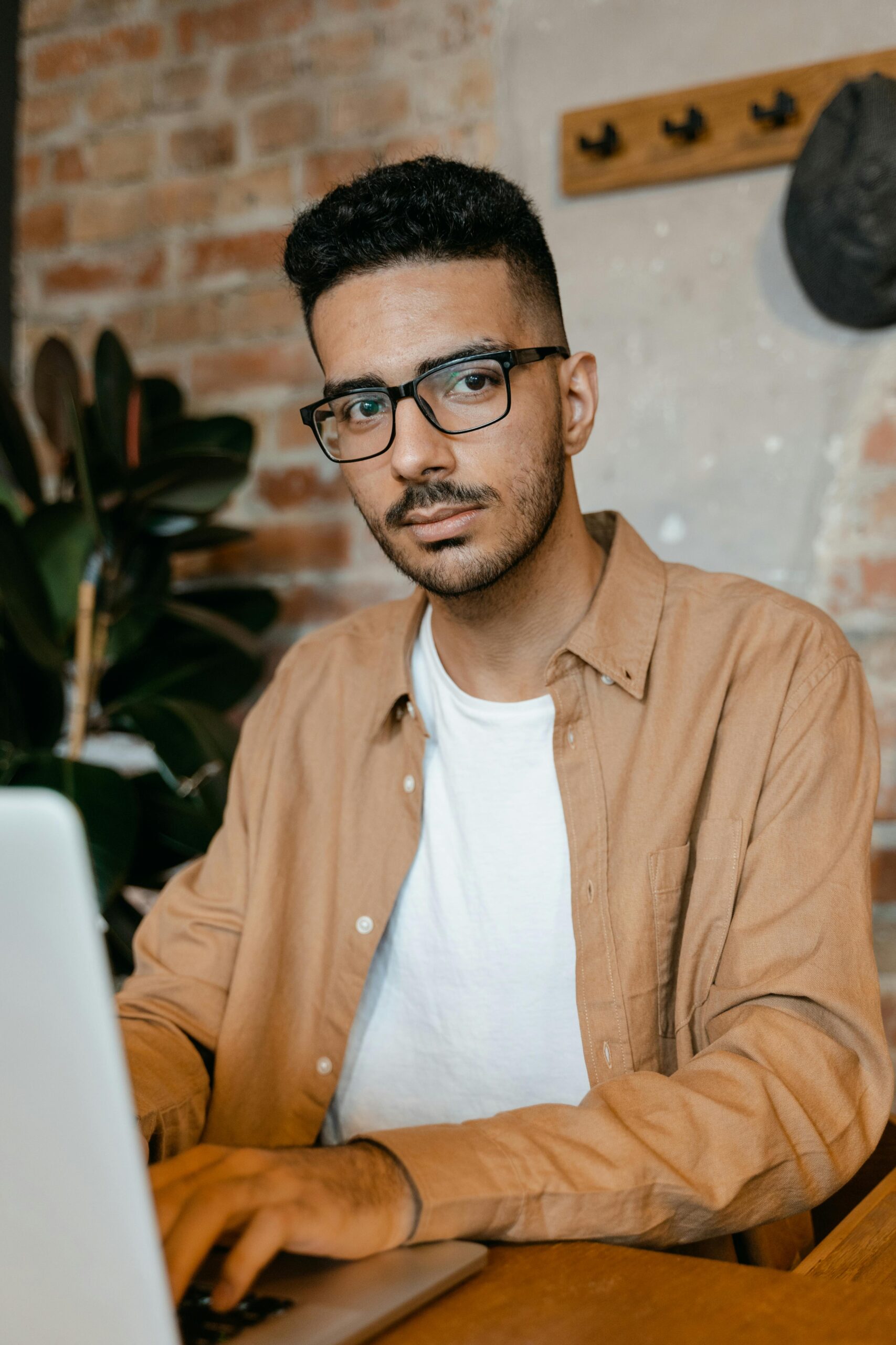 Young man wearing glasses and brown shirt working on a laptop in a stylish brick interior.